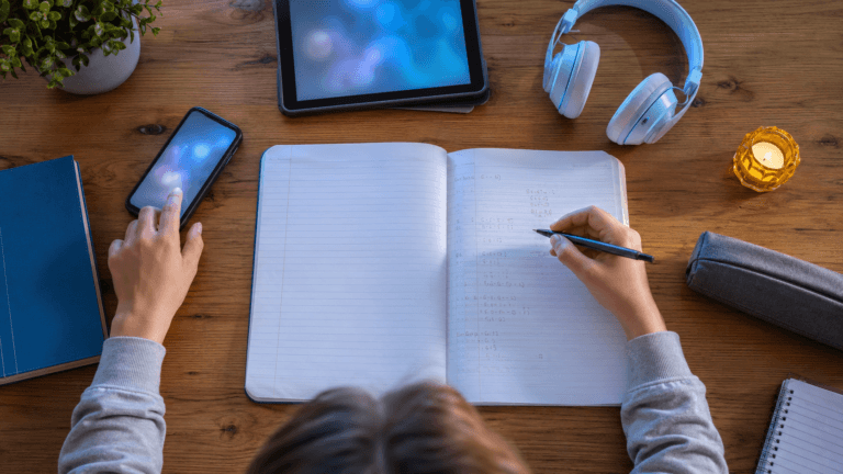 Uczeń odrabia lekcje przy biurku, spoglądając w stronę leżącego obok smartfona